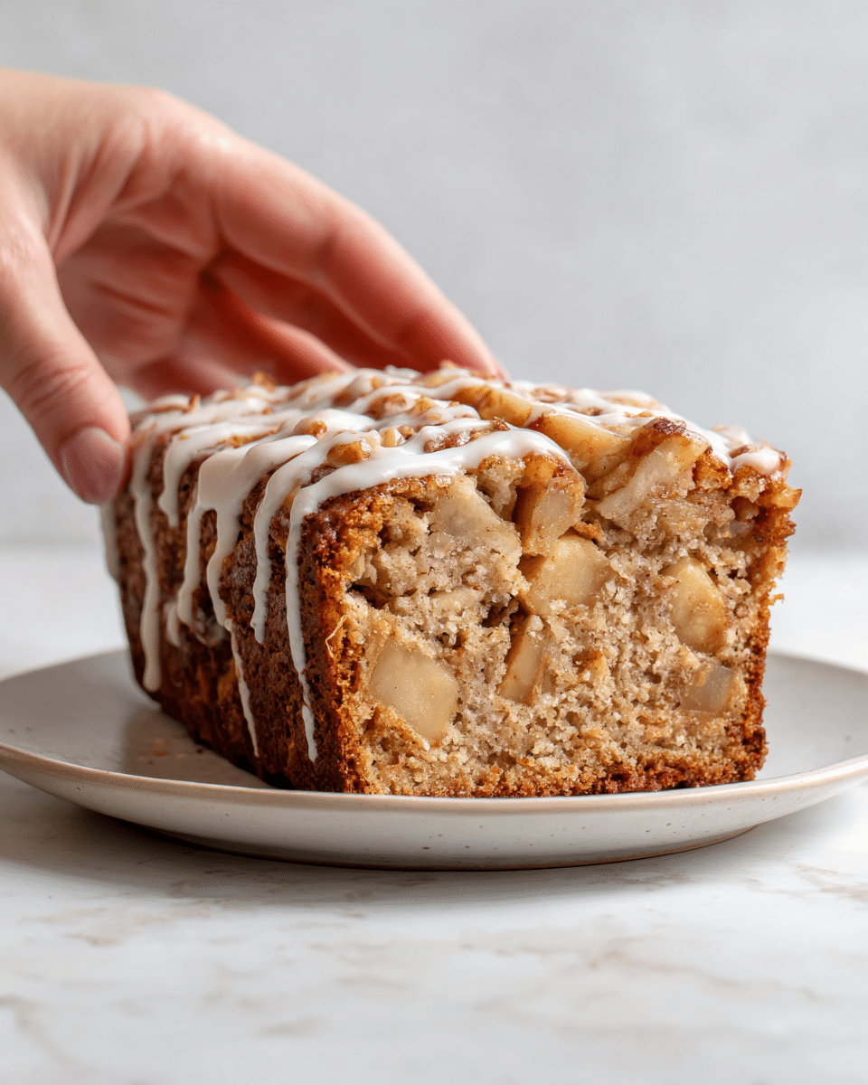 The image shows a close-up of a spiced apple fritter bread slice with a thick, textured crust on top that is golden brown with visible chunks of apple and cinnamon specks. The inside of the bread is soft and light brown with apple pieces spread throughout, showing a moist and dense texture. A smooth drizzle of white icing covers the top of the bread, contrasting with the warm colors of the bread and highlighting the bread's detailed surface. The bread is placed on a white plate sitting on a white marbled surface. A woman's hand is gently holding the plate on the side. Photo taken with an iphone --ar 4:5 --v 7