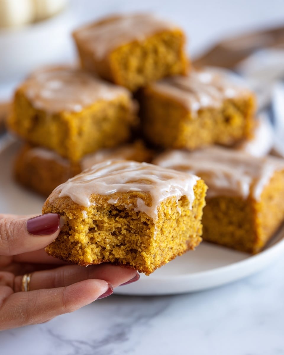 A close-up image shows a piece of soft pumpkin protein cornbread with a golden yellow crumb and a slightly dense texture, held gently by a woman's hand. The top layer of the cornbread is coated with a smooth layer of light brown sugar-free cinnamon icing, giving a glossy finish. In the background, a white marbled surface holds more square pieces of the cornbread, each topped evenly with the same cinnamon icing. The lighting highlights the moistness and texture of the cornbread, making it look inviting and fresh. Photo taken with an iphone --ar 4:5 --v 7